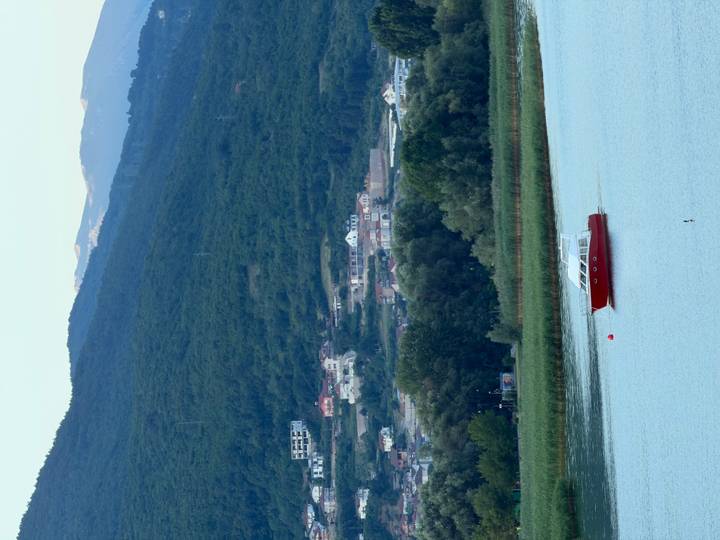 A lone red boat drifts on a tranquil lake backed by forested hills and distant mountains.