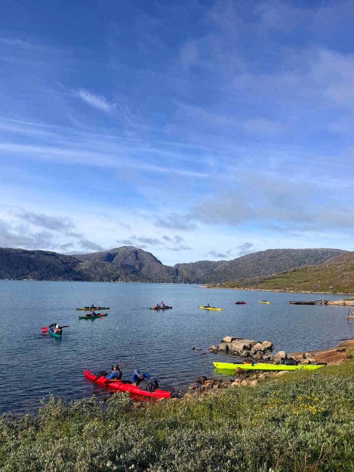 Colorful kayaks spread across a calm fjord surrounded by rugged granite peaks under a blue sky.