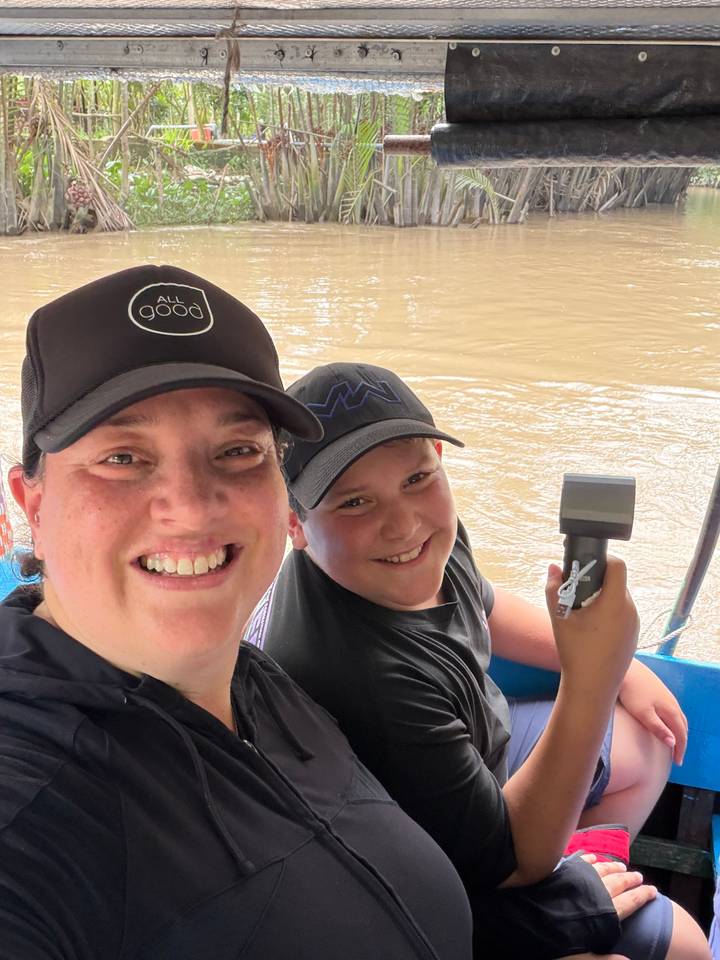 Mother and son on a boat cruising a muddy Mekong waterway, both wearing black caps