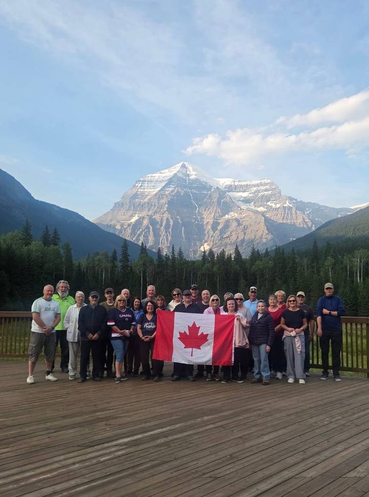 Large tour group holding a Canadian flag with a snow-capped mountain towering behind a forested valley.