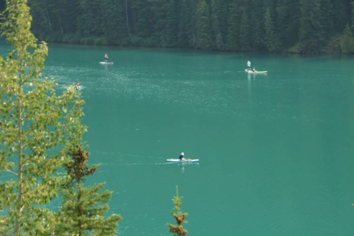 Paddle boarders and kayakers glide across a turquoise alpine lake surrounded by dense evergreen trees.
