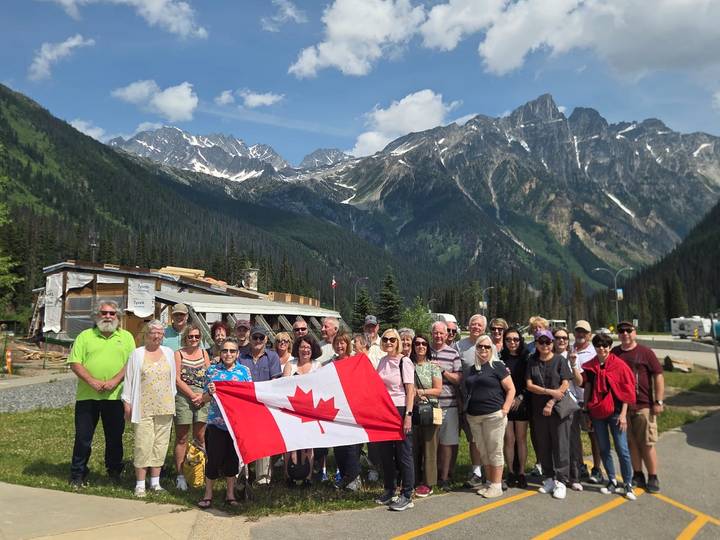 Tour group posing with a Canadian flag in front of dramatic snow-capped peaks and green valley.
