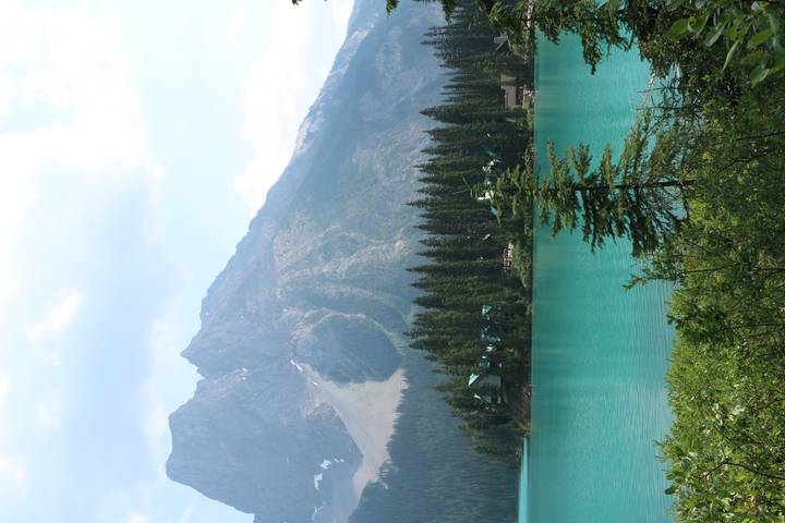Turquoise alpine lake framed by conifer trees with a rugged mountain backdrop under partly cloudy skies.
