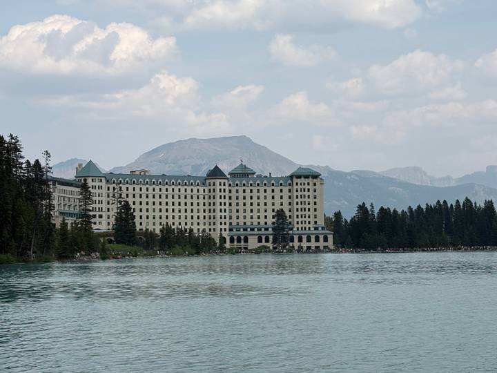 Historic grand hotel lining the edge of a turquoise mountain lake beneath hazy peaks.