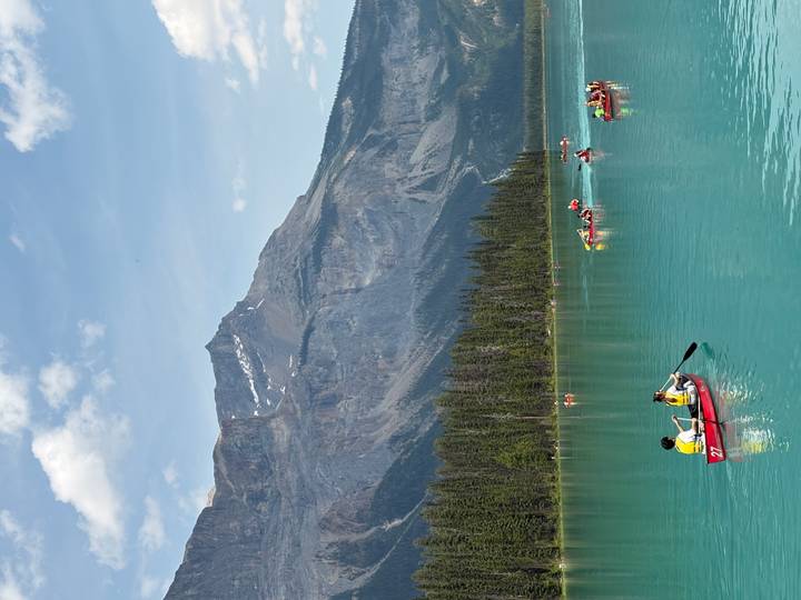 Colorful canoes with paddlers dot a bright turquoise lake surrounded by forested mountains.