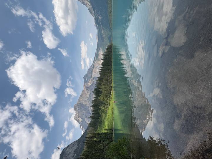 Crystal-clear alpine lake mirroring green forests and towering rocky peaks under a bright sky.