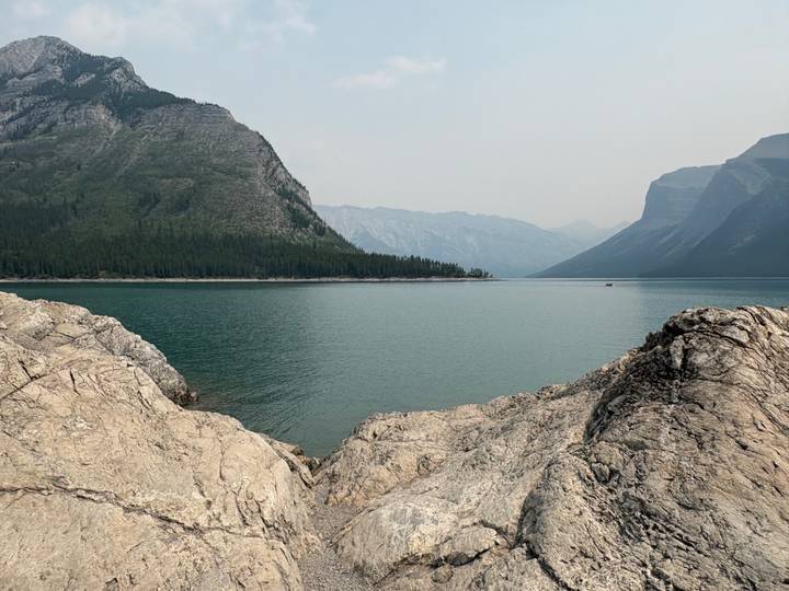 Rocky shoreline overlooking a tranquil green lake surrounded by hazy mountain ridges.