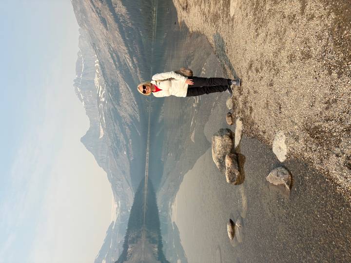 Traveler posing beside a calm mountain lake with alpine peaks mirrored in the still water.