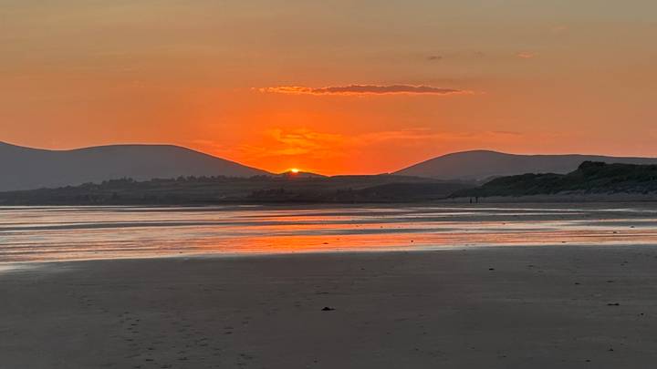 Orange sun setting behind gentle hills reflecting warm light across a wide sandy beach.