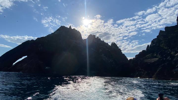 Rugged sea stacks and arch of Skellig Michael silhouetted against bright sun and scattered clouds.