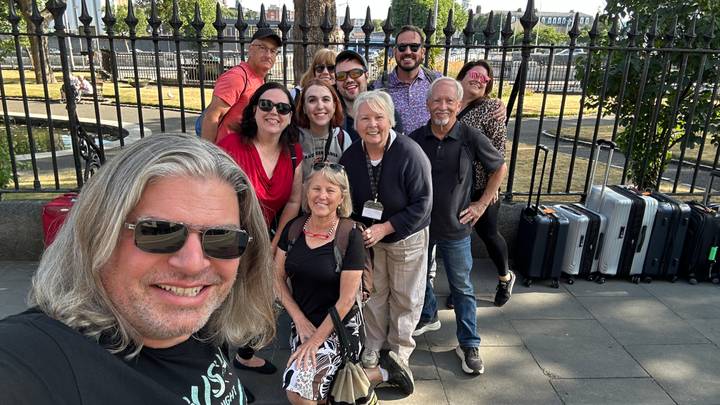 Smiling tour group posing with their suitcases on a sunny city sidewalk.