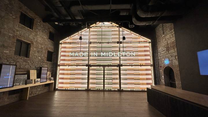 Illuminated wall of whiskey bottles spelling 'MADE IN MIDLETON' inside a historic stone distillery.