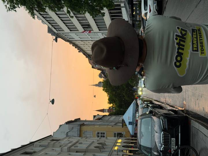 Traveller in a wide-brimmed hat photographs a pastel sunset down a European city street lined with cars.