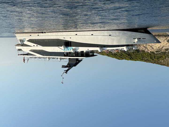 Sleek white-and-black motor yacht anchored off a rocky Mediterranean coastline under clear skies.