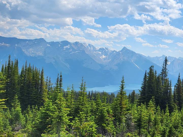 Panoramic alpine view of jagged peaks above turquoise lake and dense conifer forest.