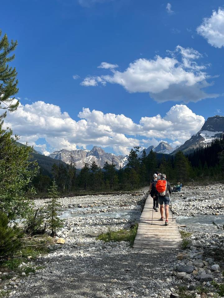 Hikers cross a wooden footbridge toward snow-capped mountains under a sky of fluffy clouds.