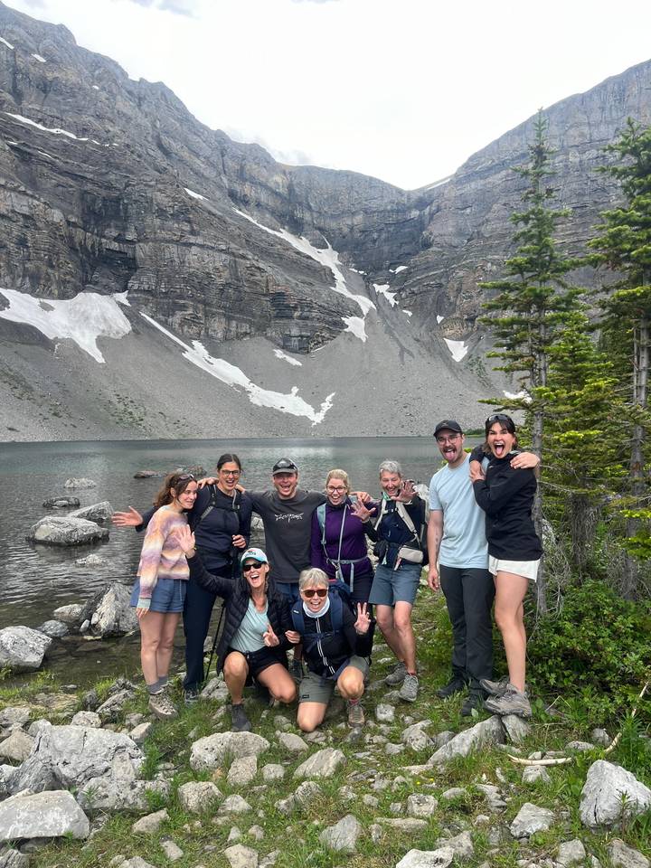 Happy hiking group poses by an alpine lake beneath steep rocky cliffs and lingering snow.