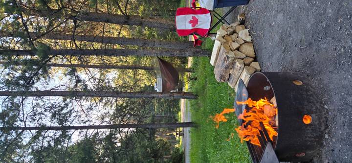 Evening campsite with crackling fire pit, stacked wood, hammock and a chair with Canadian flag.