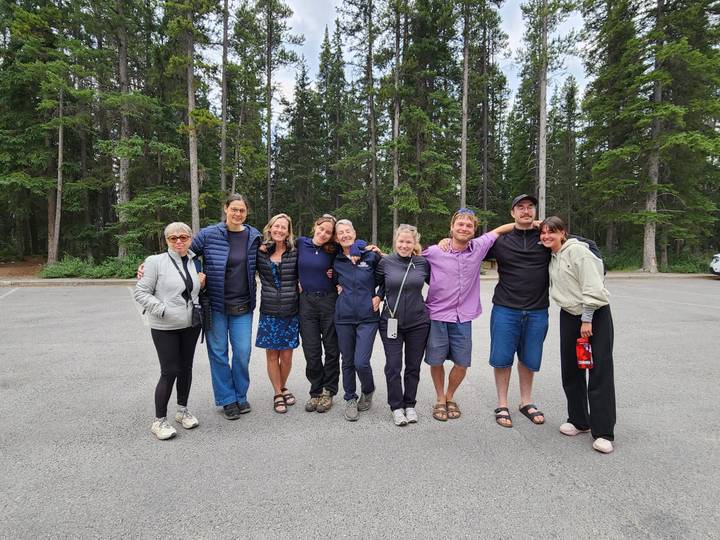 Travel group poses arm-in-arm in parking lot with tall pine forest backdrop.