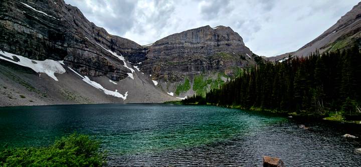 Clear emerald alpine lake beneath towering grey cliffs and scattered snow patches.
