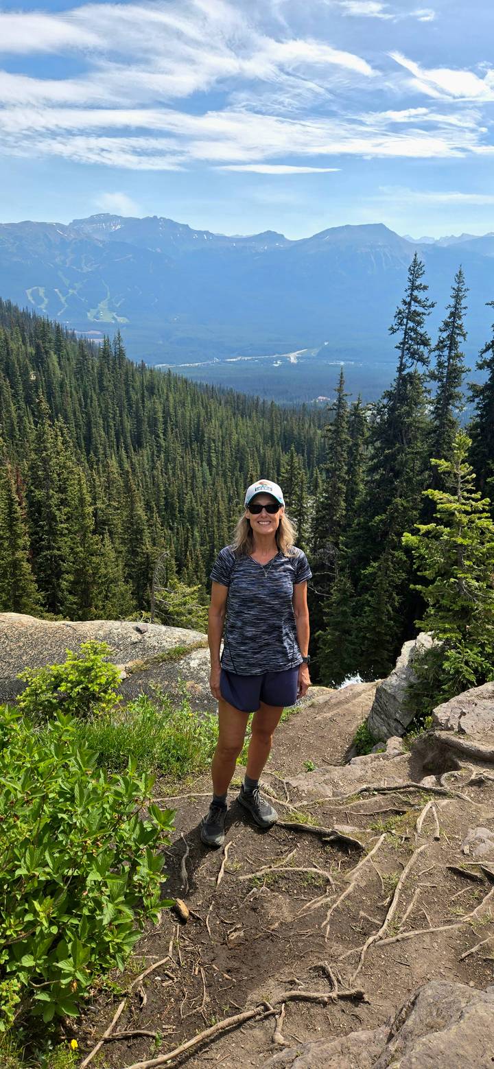 Smiling woman in cap stands on rocky outcrop with dense alpine forest stretching behind.
