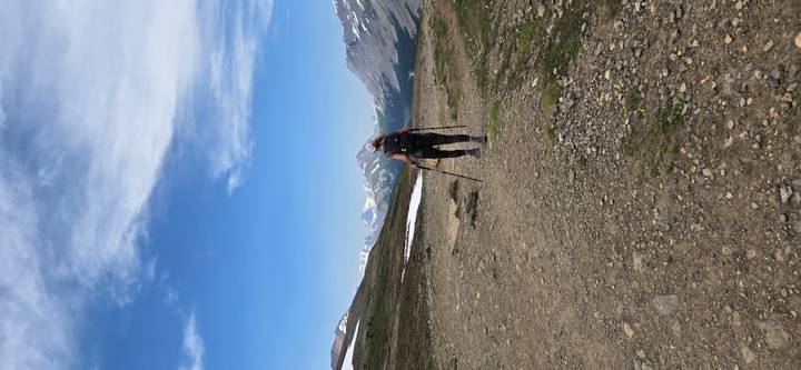 Backpack-laden hiker with trekking poles walks along a high alpine trail toward snow-capped peaks.