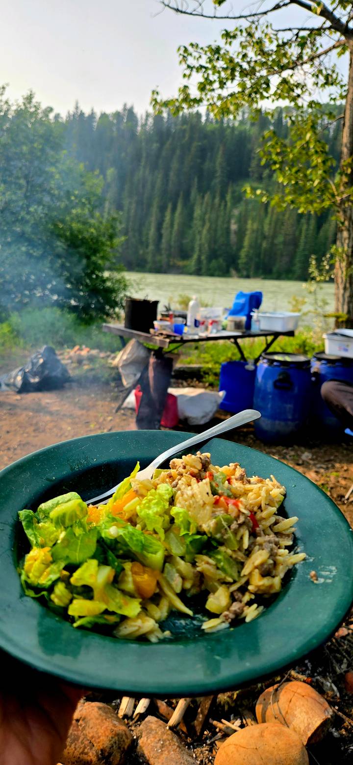 Close-up of a camp meal in a bowl with a blurry background of cooking gear set beside a lake.