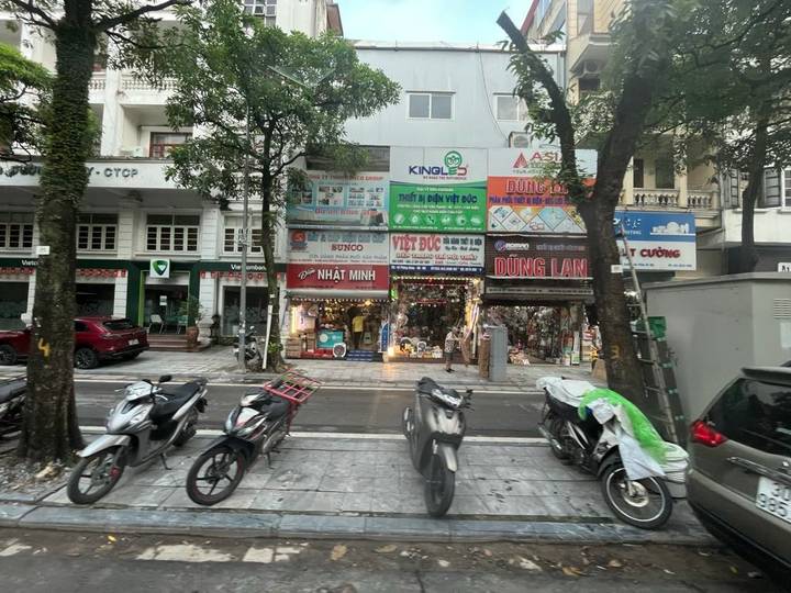 Busy Hanoi street with motorbikes parked in front of small multi-story shopfronts and billboards.