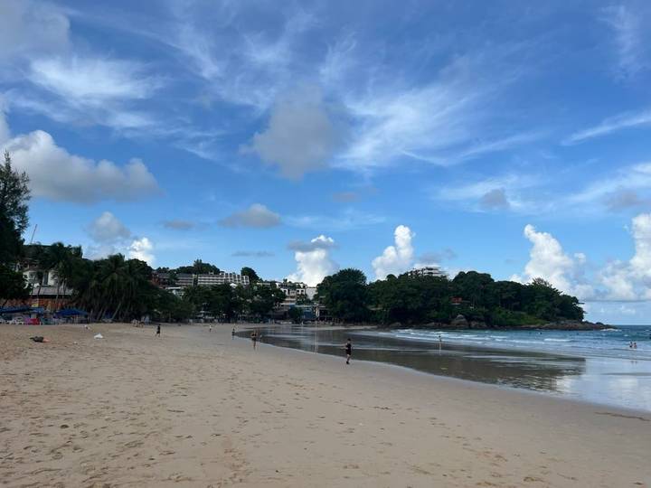 Wide sandy beach with a few walkers, backed by tropical trees and low buildings under scattered clouds.