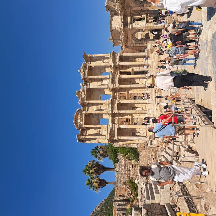 Crowds explore the grand façade of the ancient Celsus Library in Ephesus under a deep blue sky.