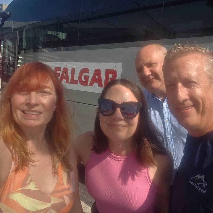 Casual selfie of four travellers smiling in front of a tour coach on a sunny day