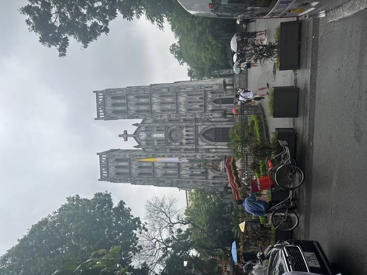 Gothic-style cathedral facade in a city square with rickshaw, pedestrians and umbrellas on a grey day