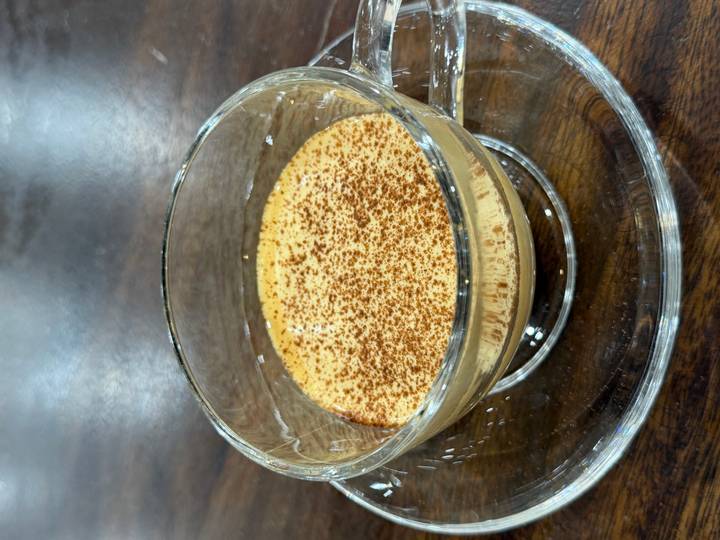 Glass cup of creamy Vietnamese egg coffee topped with cocoa powder on a clear saucer.