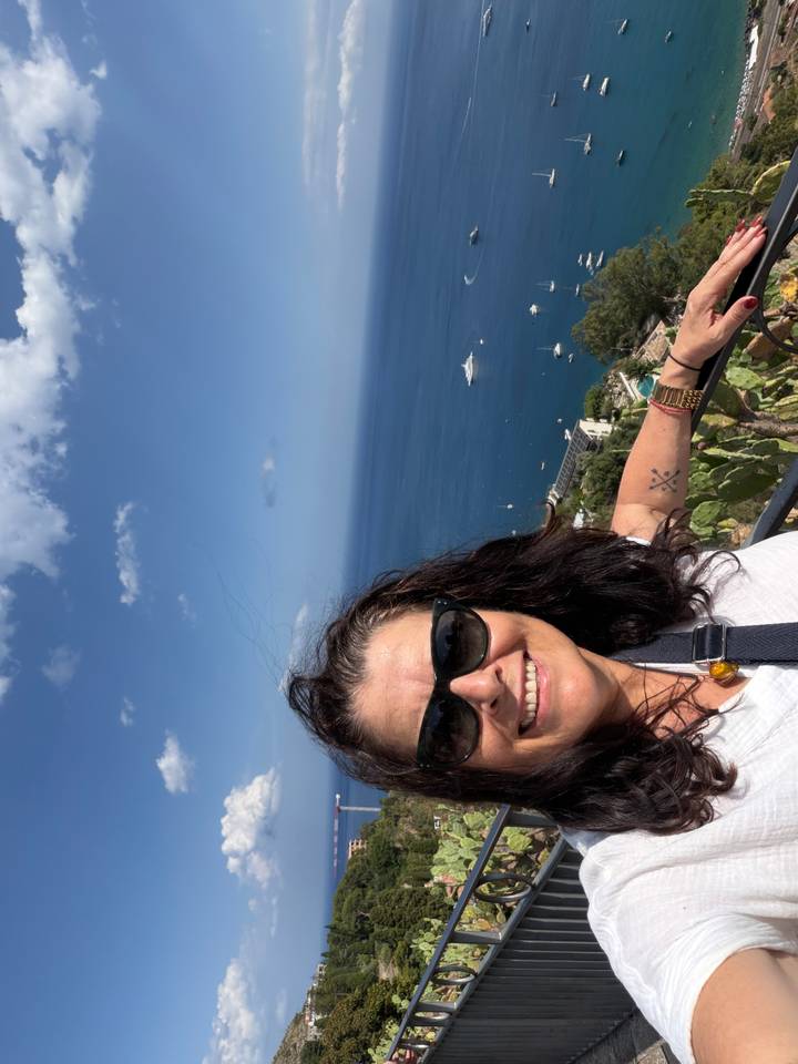 Selfie of a smiling woman wearing sunglasses at a high seaside lookout with deep-blue Mediterranean waters and yachts below