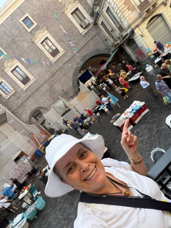 Overhead scene of a busy outdoor market square with stalls and shoppers; a woman in a white hat peeks into frame pointing downward
