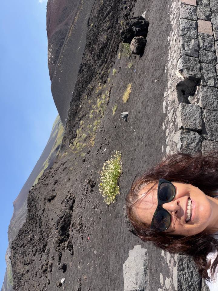 Face of a woman in sunglasses with rugged black lava slopes and flowering shrubs rising toward Mount Etna behind
