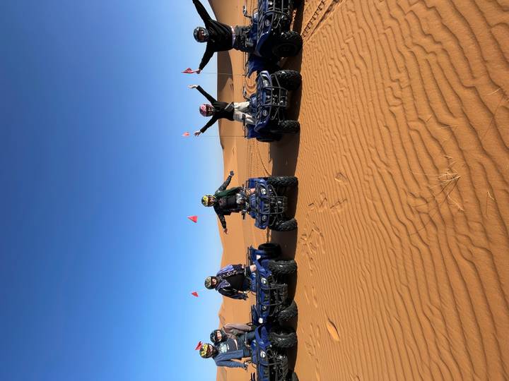 Four riders celebrating on quad bikes atop golden desert dunes under a clear sky.