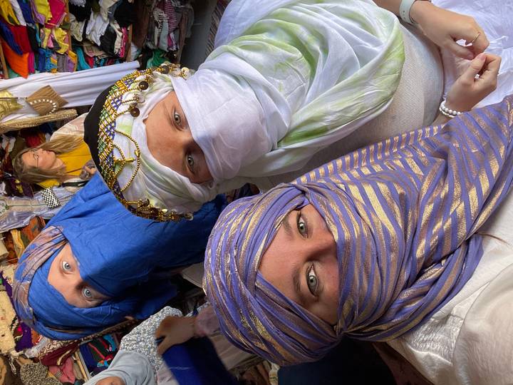 Three friends wearing vibrant desert scarves inside a colorful market stall.