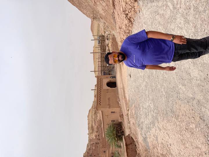 Traveller standing on a dusty track outside an earthen kasbah structure on an overcast desert day.
