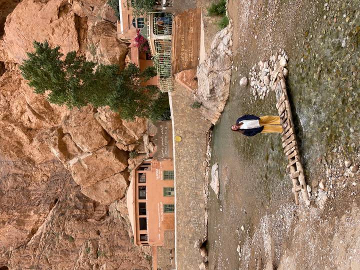 Traveller standing on a small wooden footbridge over a stream beneath towering ochre cliffs.