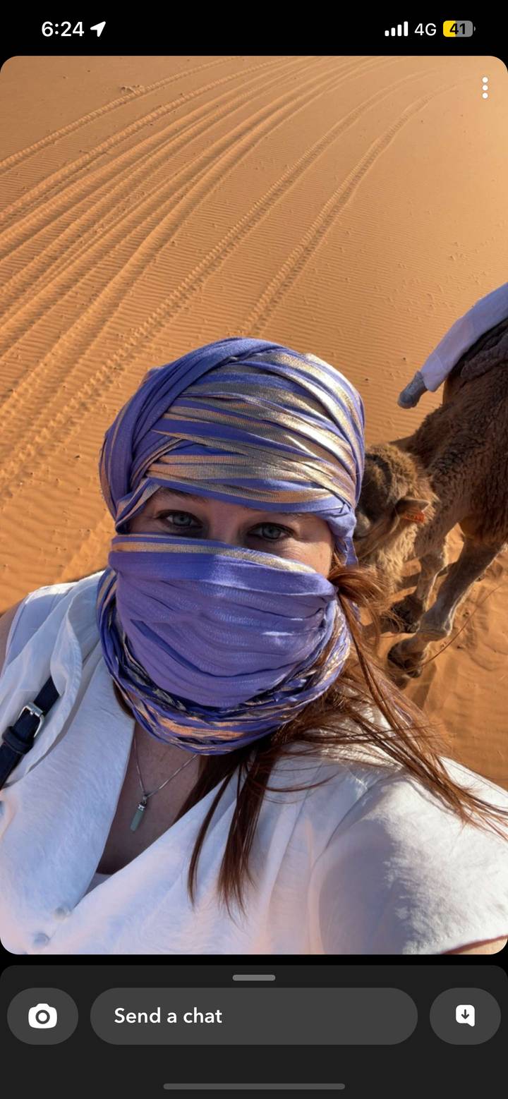 Close-up of a traveller with purple headscarf in desert with camel partially visible behind.