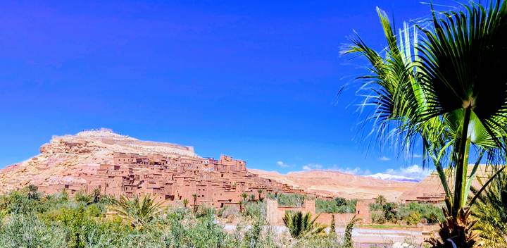 Ancient mud-brick citadel of Ait Benhaddou rises above a lush oasis under a deep blue sky.