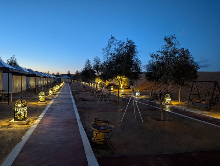 Evening view of a desert camp walkway lined with illuminated Moroccan lanterns and tents beside sand dunes