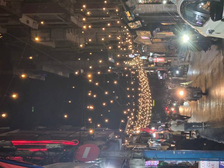 Bustling night street decorated with hanging string lights and traffic of scooters and pedestrians.