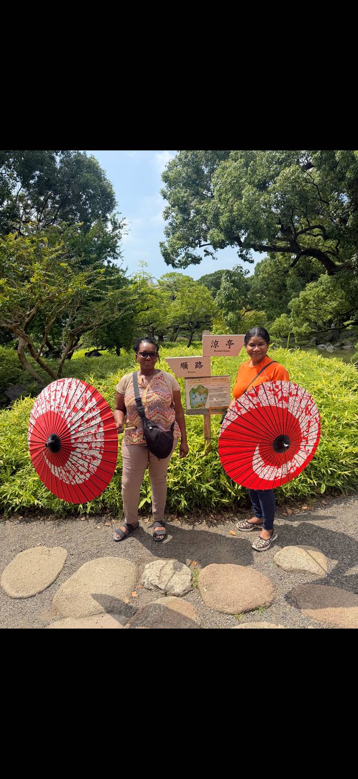 Two women holding traditional red parasols in a lush Japanese garden.