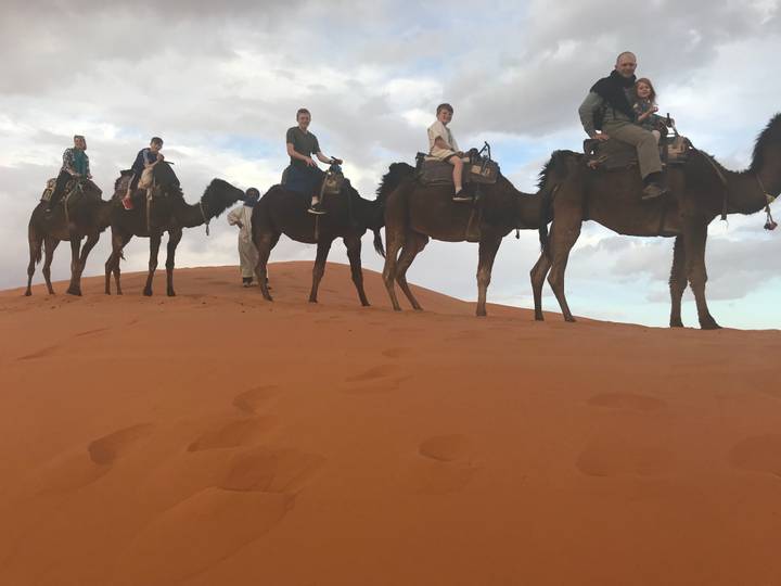 Line of tourists riding camels across orange Sahara dunes beneath a cloudy sky.