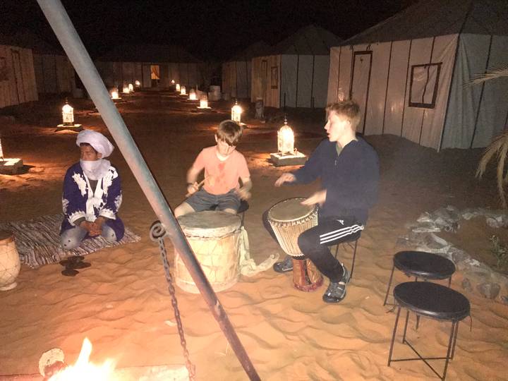 Three boys play drums around a campfire line of lanterns in a desert camp at night.