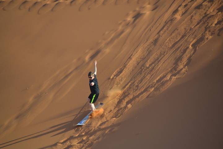 Boy sandboards down a steep dune leaving a trail of orange dust behind him.