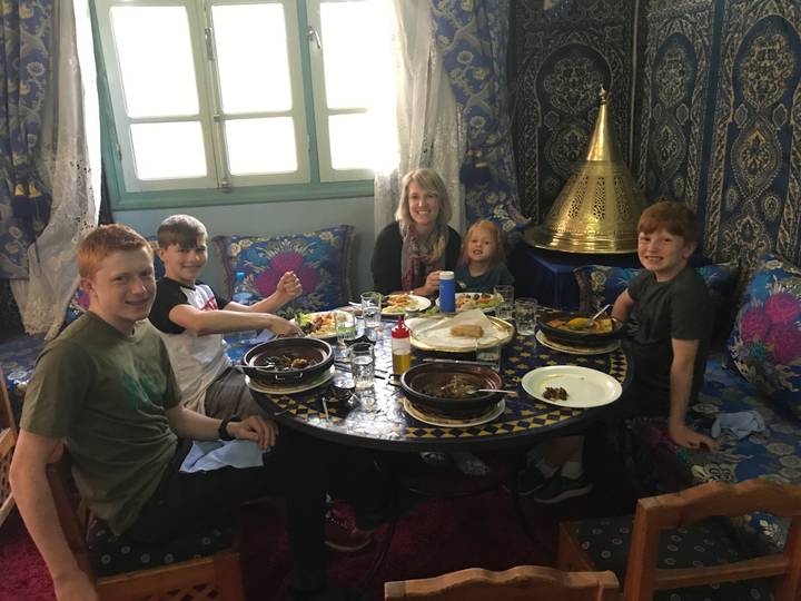 Family enjoys traditional Moroccan dishes around a round mosaic table in a richly decorated room.