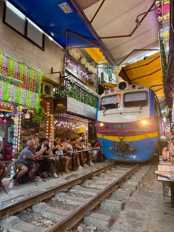 A train squeezes past a lively street-side café while diners watch and photograph the spectacle.